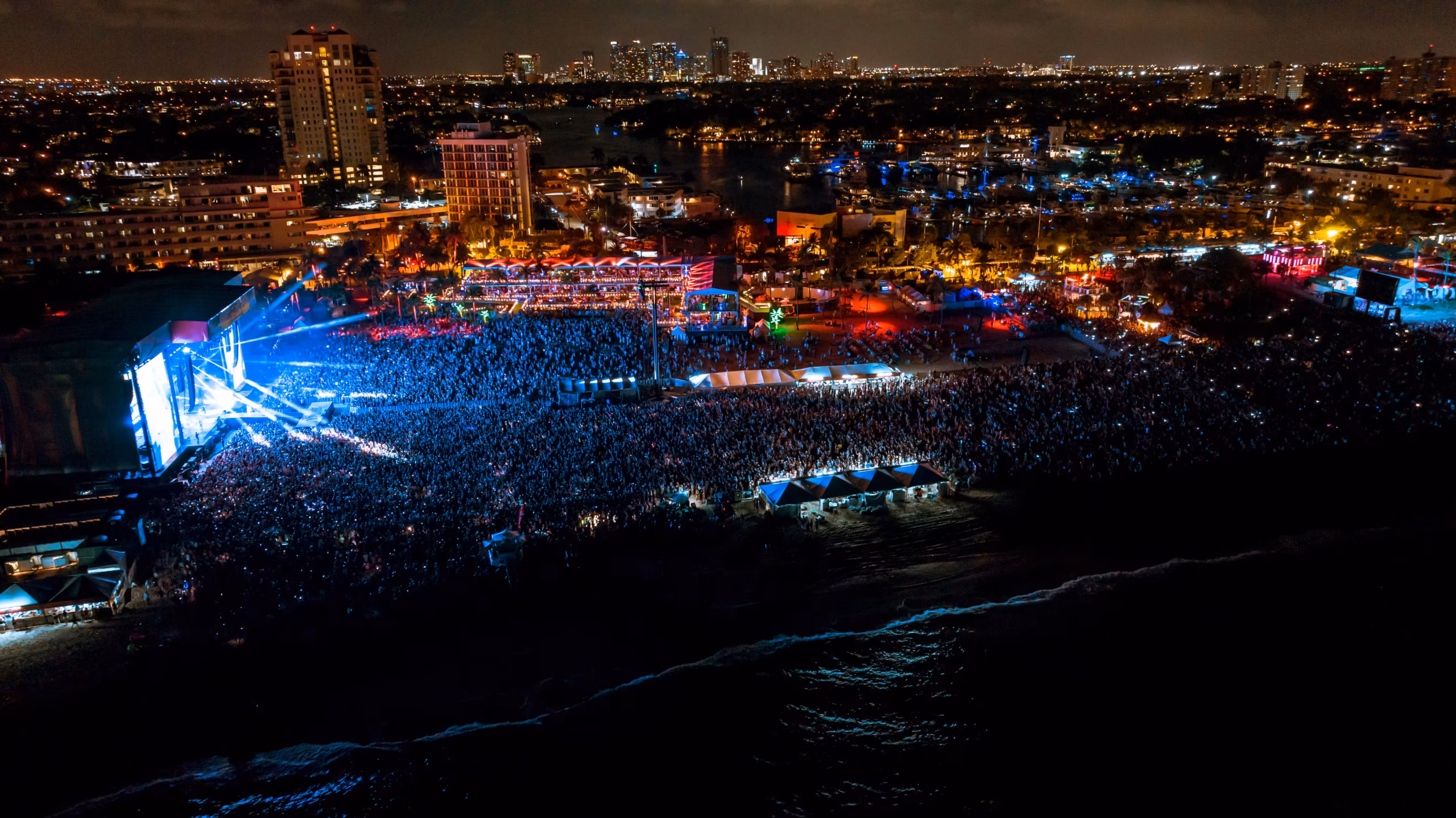 Aerial drone view of Tortuga Music Festival at night with blue stage lights illuminating the crowd on Fort Lauderdale Beach with the city skyline in the background
